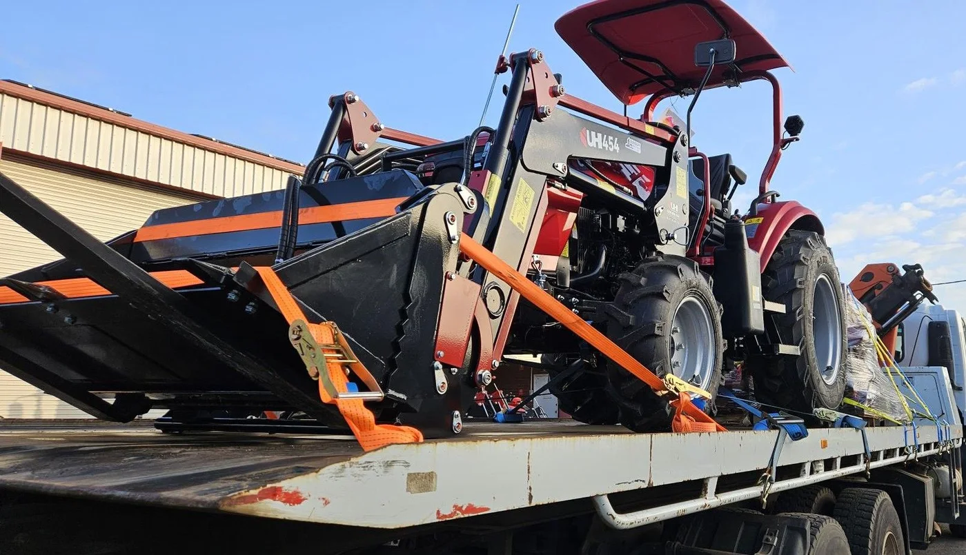 Compact tractor with front loader secured on a flatbed for transport.