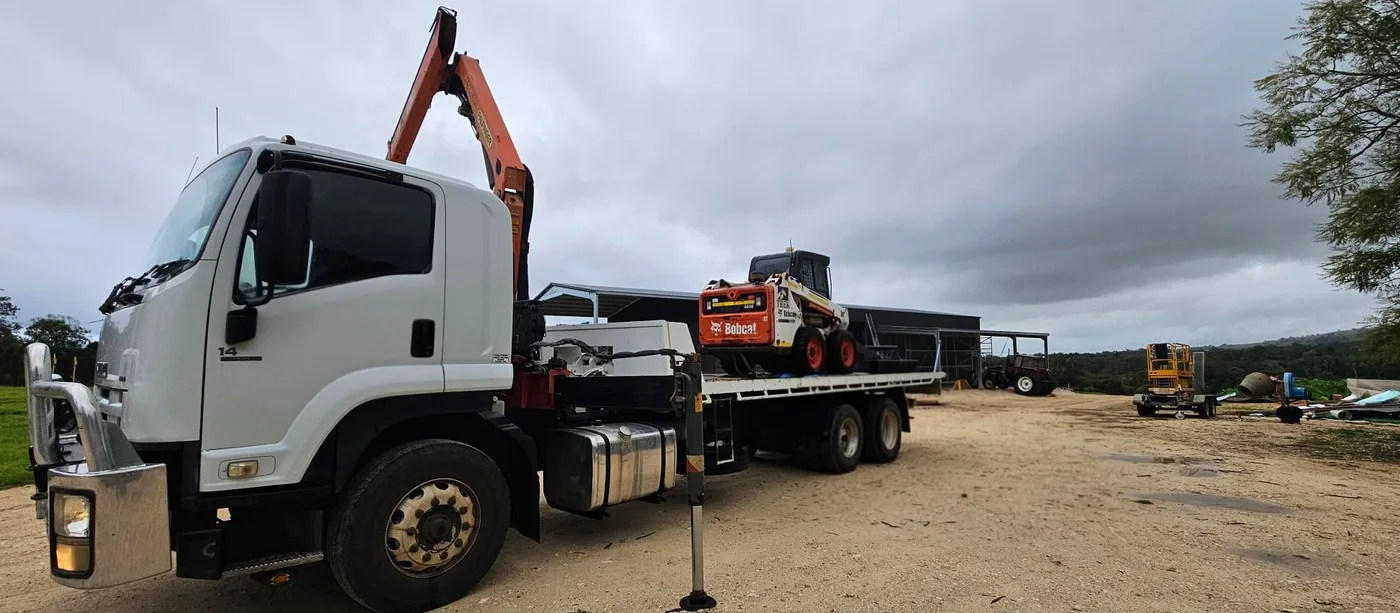 White hiab truck loaded with a Bobcat skid steer at a rural property.