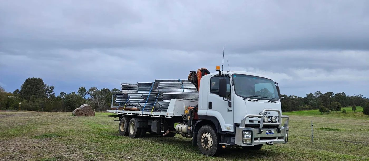 Hiab truck transporting galvanized pipe and fencing materials through a paddock.