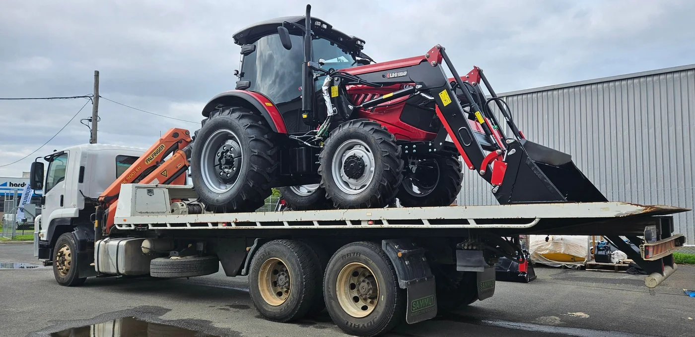 Red tractor loaded on a flatbed truck outside an industrial shed.