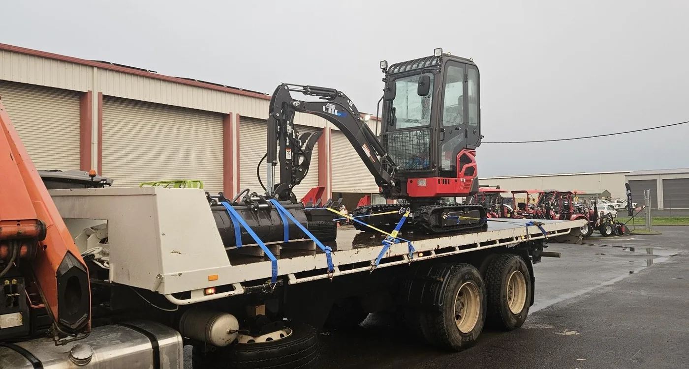 Mini excavator secured on a flatbed outside an equipment depot.