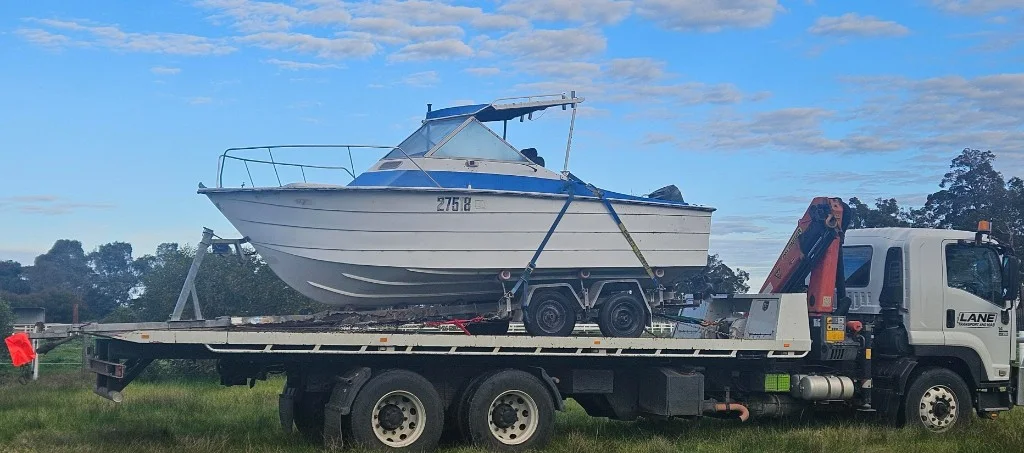 Lane Transport hiab truck carrying a white motorboat on a trailer across a South West paddock.